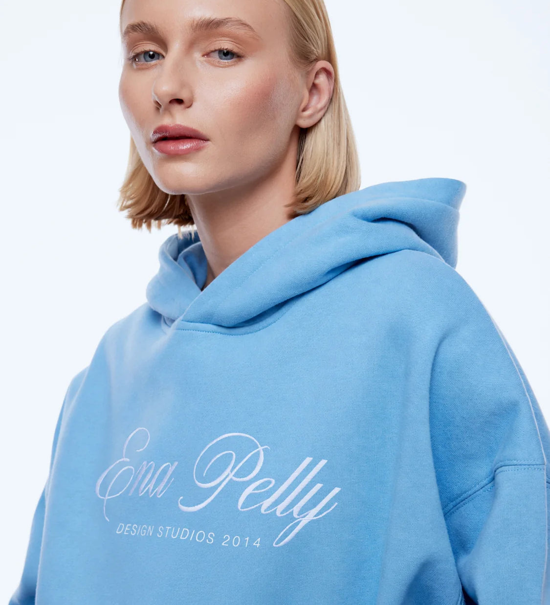 A blonde woman wearing a light blue pullover hoodie with 'Ena Pelly' and 'DESIGN STUDIOS 2014' printed in white script on the front, set against a plain white studio background.