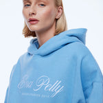A blonde woman wearing a light blue pullover hoodie with 'Ena Pelly' and 'DESIGN STUDIOS 2014' printed in white script on the front, set against a plain white studio background.