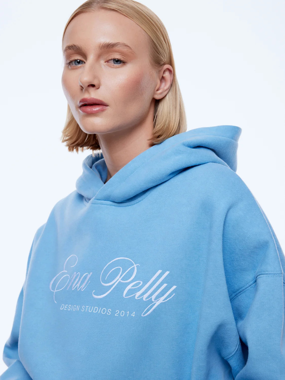 A blonde woman wearing a light blue pullover hoodie with 'Ena Pelly' and 'DESIGN STUDIOS 2014' printed in white script on the front, set against a plain white studio background.