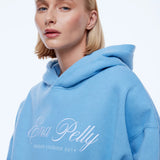 A blonde woman wearing a light blue pullover hoodie with 'Ena Pelly' and 'DESIGN STUDIOS 2014' printed in white script on the front, set against a plain white studio background.