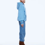 A model is shown from the back wearing a light blue oversized hoodie, blue jeans, and brown heeled ankle boots against a plain white studio background.