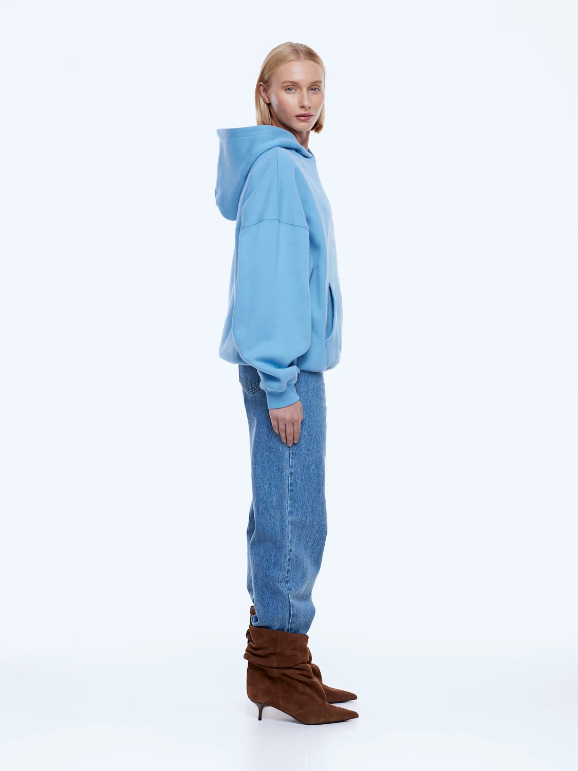 A model is shown from the back wearing a light blue oversized hoodie, blue jeans, and brown heeled ankle boots against a plain white studio background.