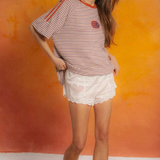Woman wearing a striped shirt and white shorts against an orange wall.