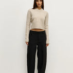 Woman wearing a beige sweater and black pants against a white background