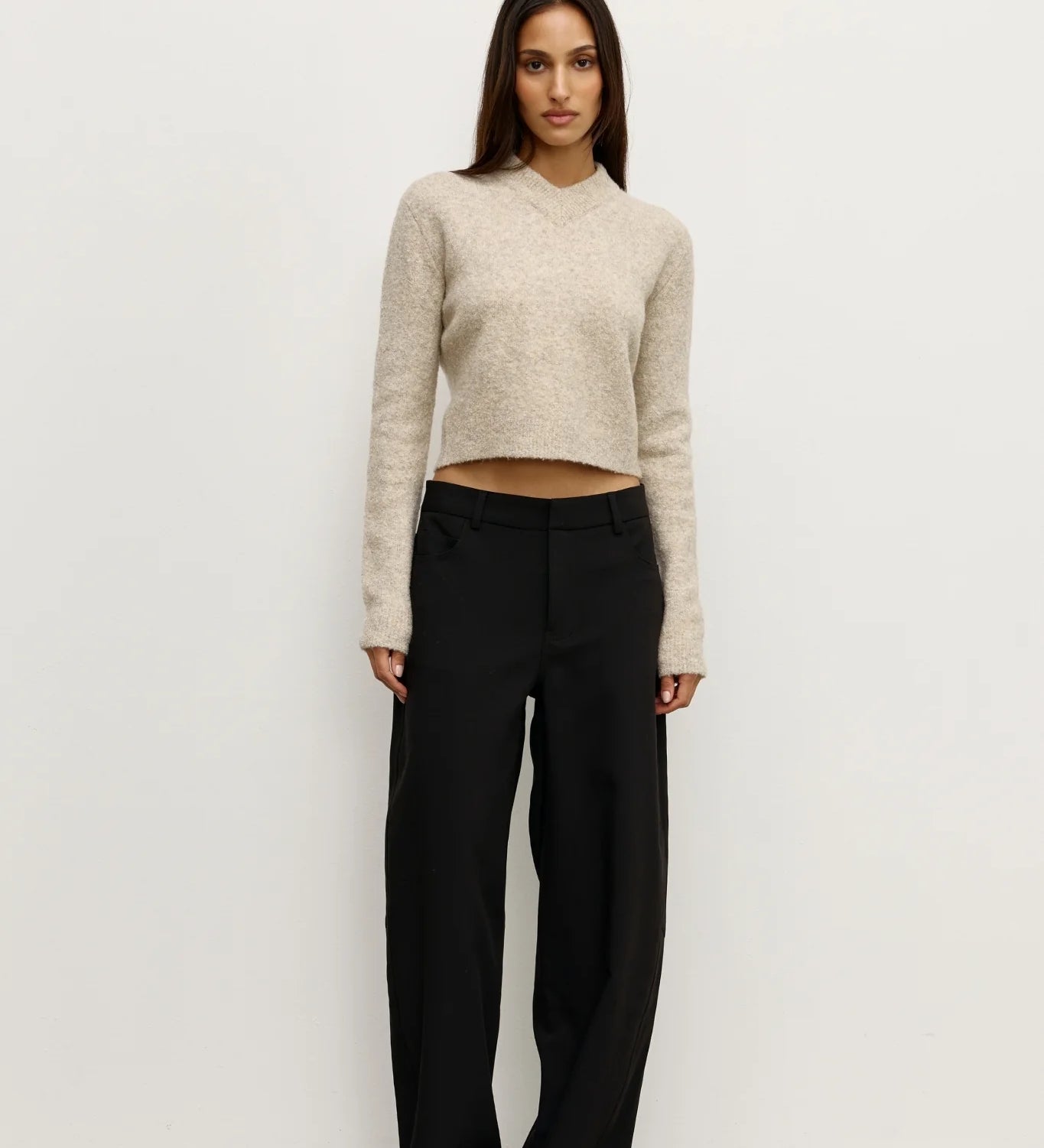 Woman wearing a beige sweater and black pants against a white background