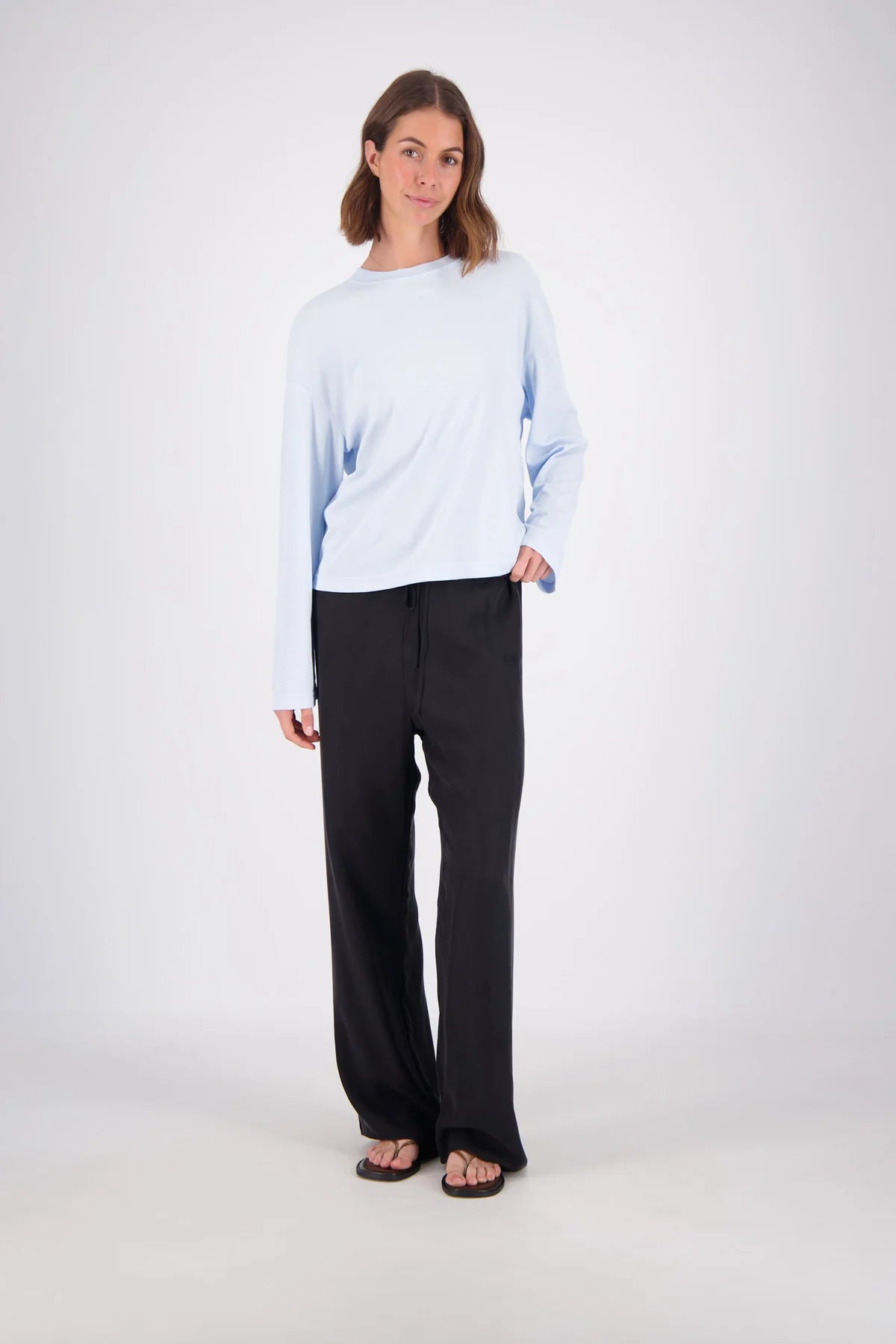 A woman modeling a light blue long-sleeve crewneck top and black wide-leg pants against a plain light gray studio background.