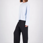 A woman modeling a light blue long-sleeve crewneck top and black wide-leg trousers, standing against a plain white studio background.