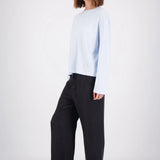 A woman modeling a light blue long-sleeve crewneck top and black wide-leg trousers, standing against a plain white studio background.