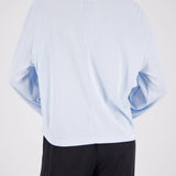 A person shown from the back wearing a light blue cropped long-sleeve top and black pants against a plain light gray studio background.