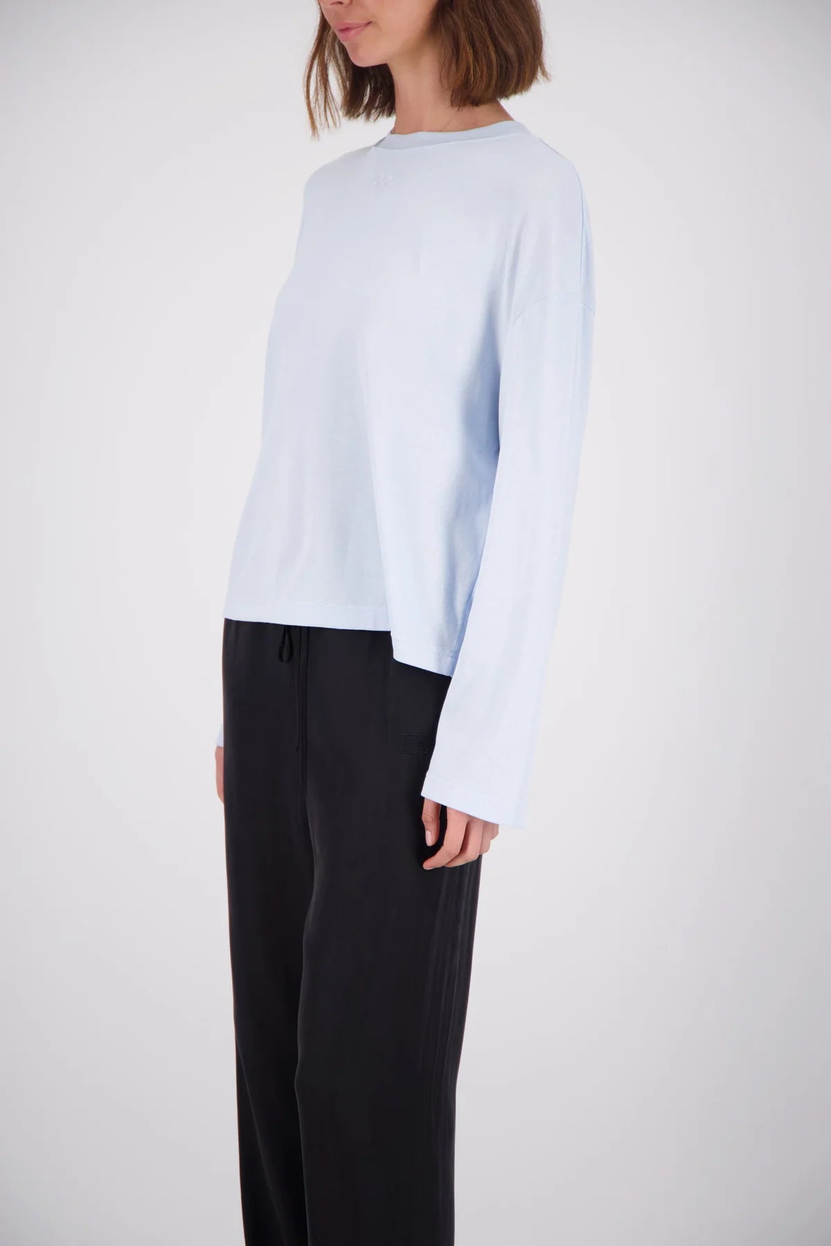 A model wearing a light blue long-sleeve knit top and black wide-leg trousers, shown from the side against a light gray studio background.