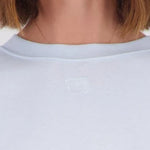 Close-up of a person wearing a light-colored crewneck top, showing the neck, chin, and hair against a plain light background.