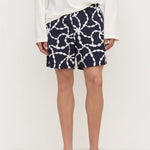 A person wearing navy shorts with a white chain-link pattern, a white long-sleeve top, and black flip-flops, standing against a plain white studio background.