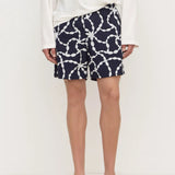 A person wearing navy shorts with a white chain-link pattern, a white long-sleeve top, and black flip-flops, standing against a plain white studio background.