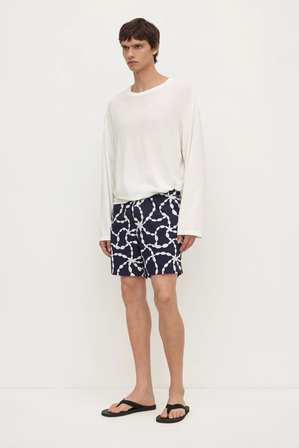A male model wearing a white long-sleeve knit top, navy blue shorts with a white chain-link pattern, and black flip-flop sandals, standing against a plain white studio background.