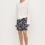 A male model wearing a white long-sleeve knit top, navy blue shorts with a white chain-link pattern, and black flip-flop sandals, standing against a plain white studio background.