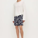 A male model wearing a white long-sleeve knit top, navy blue shorts with a white chain-link pattern, and black flip-flop sandals, standing against a plain white studio background.