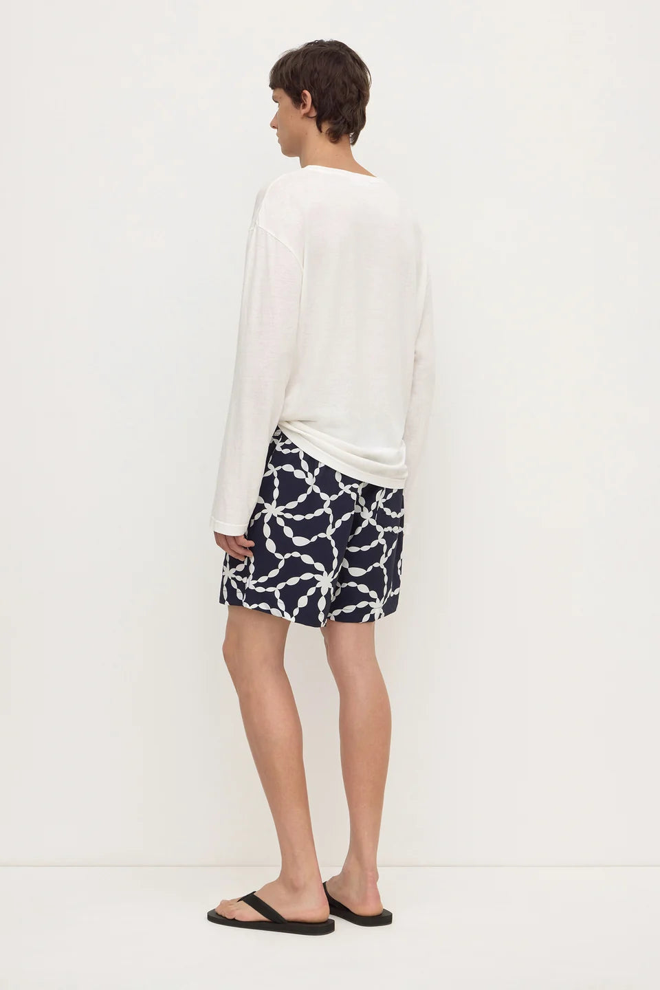 A man shown from the back wearing a white long-sleeve shirt and navy shorts with a white chain-link pattern, paired with black flip-flop sandals, against a plain white studio background.