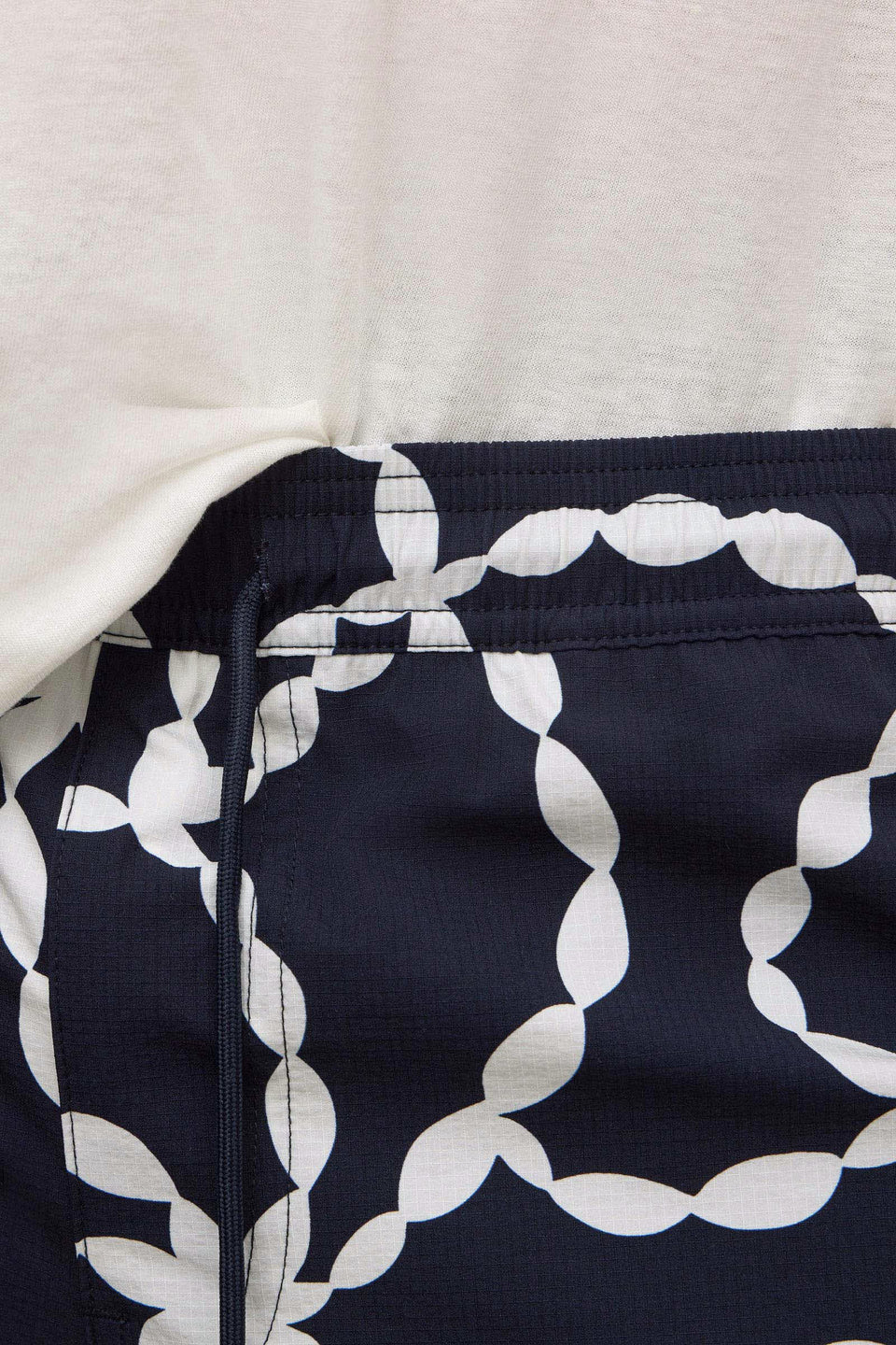 Close-up of a garment waist area showing a navy fabric with a large white chain-link pattern and a white drawstring, paired with a white top.