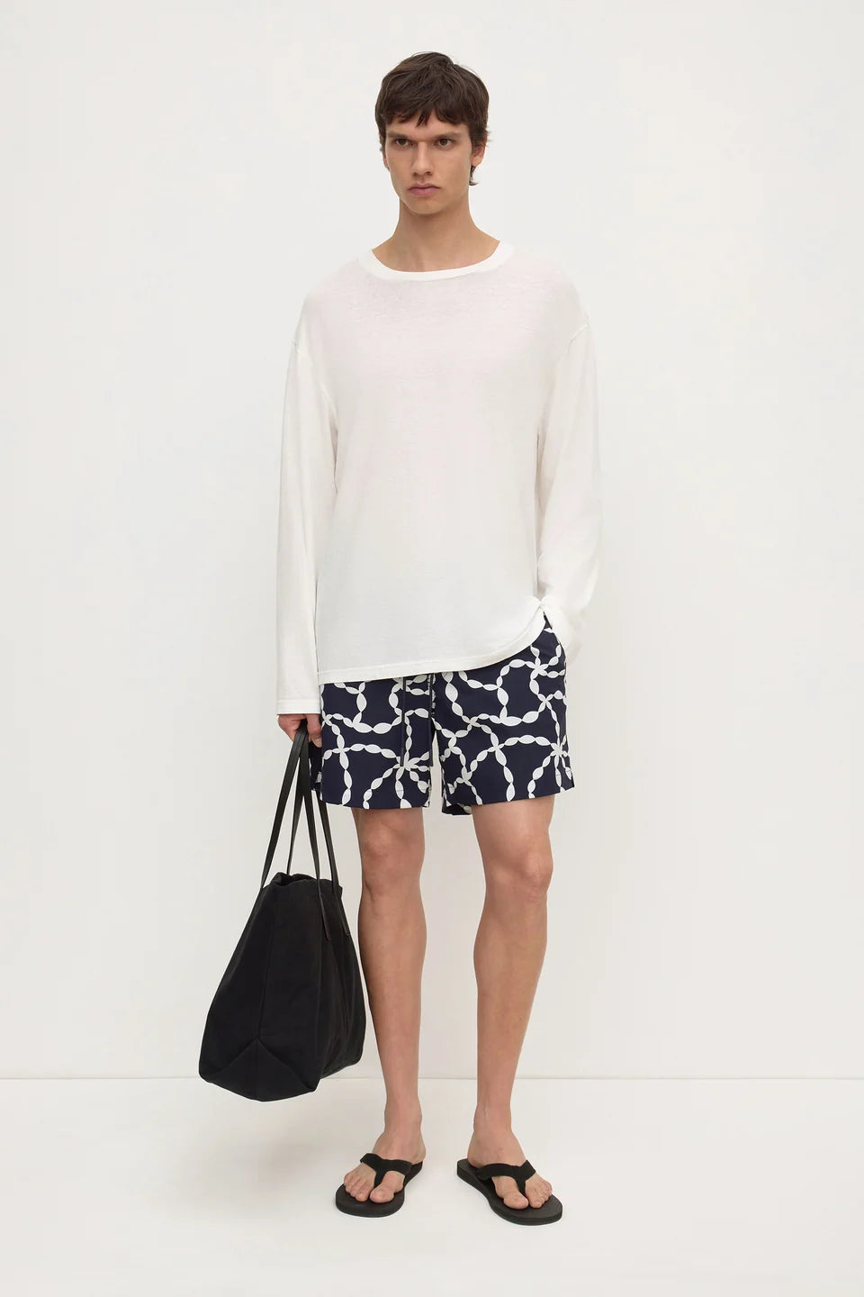 A man modeling a white long-sleeve crewneck knit top, paired with navy blue shorts featuring a white chain-link pattern, black sandals, and holding a black tote bag against a plain white studio background.
