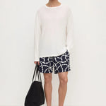 A man modeling a white long-sleeve crewneck knit top, paired with navy blue shorts featuring a white chain-link pattern, black sandals, and holding a black tote bag against a plain white studio background.