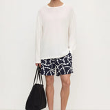 A man modeling a white long-sleeve crewneck knit top, paired with navy blue shorts featuring a white chain-link pattern, black sandals, and holding a black tote bag against a plain white studio background.