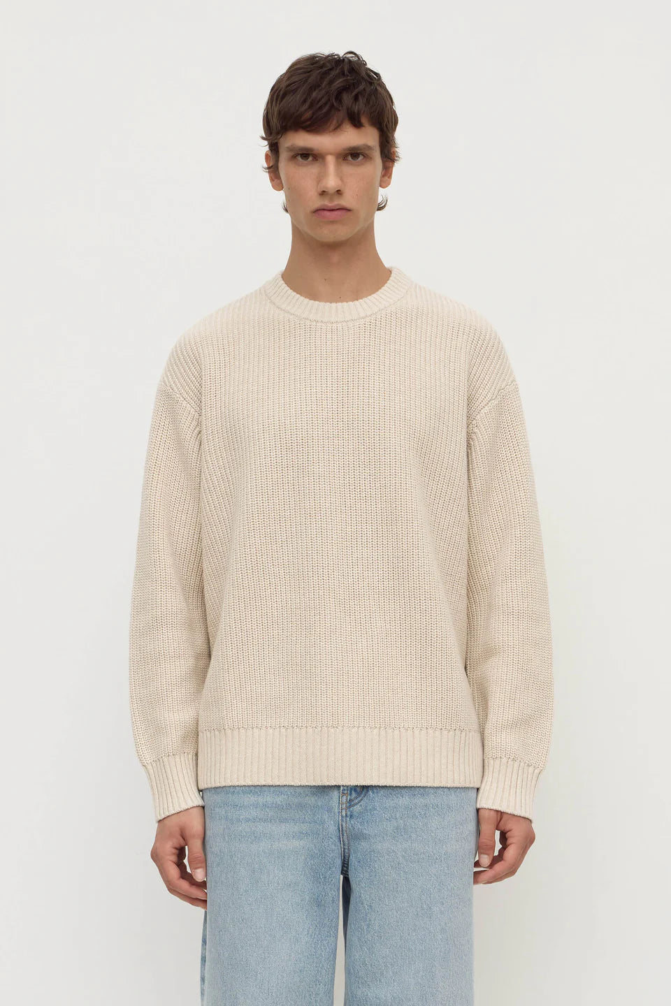 Man wearing a beige sweater and light blue jeans on a white background