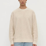 Man wearing a beige sweater and light blue jeans on a white background