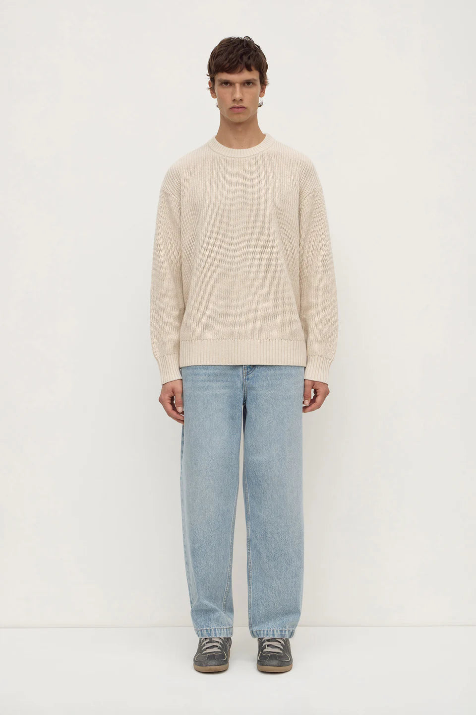 Man wearing a beige sweater and light blue jeans on a white background