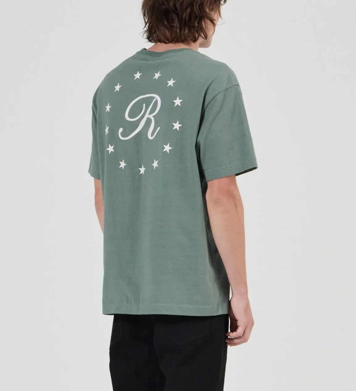 A model shown from the back wearing a sage green t-shirt with a white monogram 'R' and stars on the back, paired with black pants and gray sneakers, against a light gray studio background.