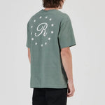 A model shown from the back wearing a sage green t-shirt with a white monogram 'R' and stars on the back, paired with black pants and gray sneakers, against a light gray studio background.