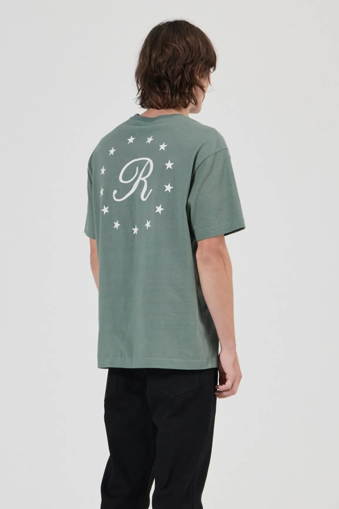 A model shown from the back wearing a sage green t-shirt with a white monogram 'R' and stars on the back, paired with black pants and gray sneakers, against a light gray studio background.