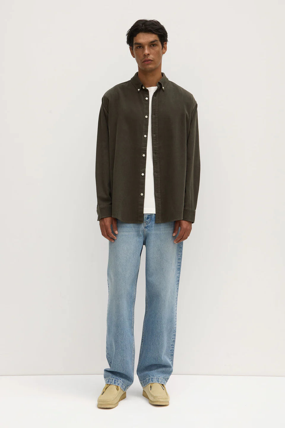 A male model wearing a long sleeve button-up shirt in sorrel color, with white contrast buttons and a U-shaped hem, paired with blue jeans and white shoes.