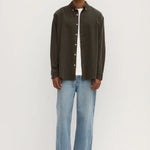 A male model wearing a long sleeve button-up shirt in sorrel color, with white contrast buttons and a U-shaped hem, paired with blue jeans and white shoes.