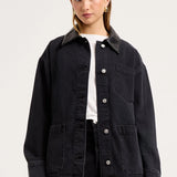 A woman modeling an oversized black denim utility jacket with a pointed collar, button-front closure, chest pocket, and large front patch pockets, worn over a white top and black pants against a light gray studio background.