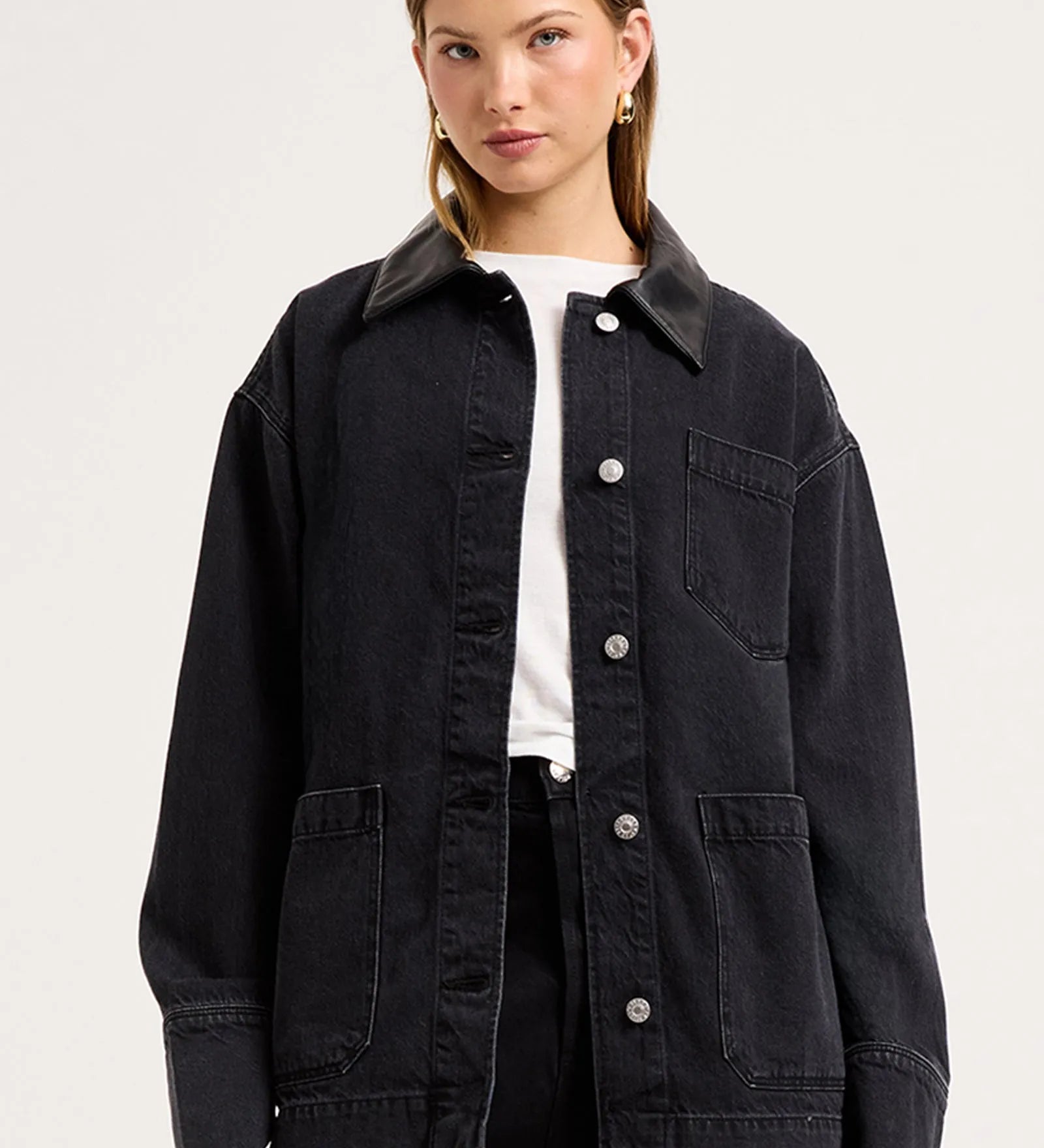 A woman modeling an oversized black denim utility jacket with a pointed collar, button-front closure, chest pocket, and large front patch pockets, worn over a white top and black pants against a light gray studio background.