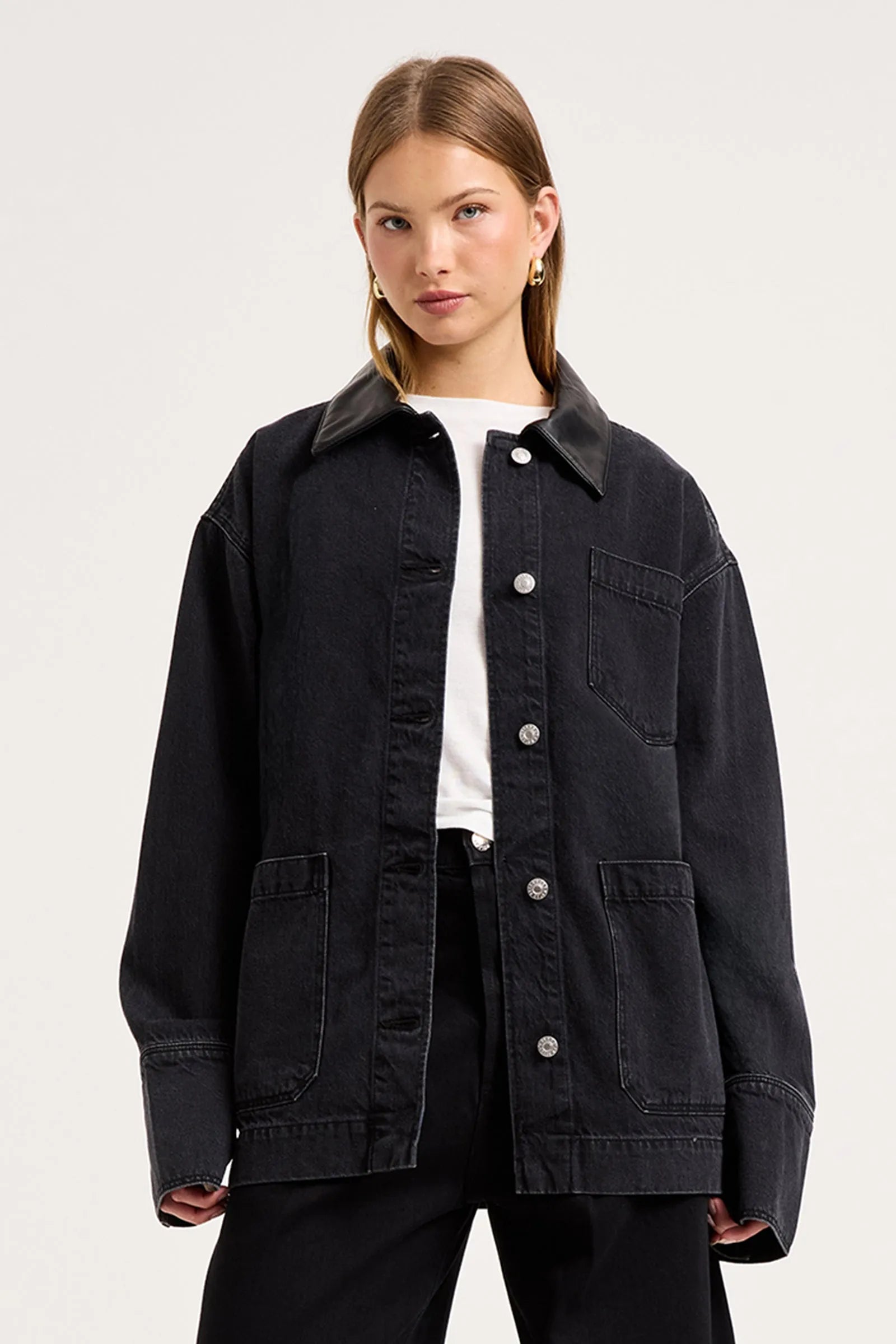 A woman modeling an oversized black denim utility jacket with a pointed collar, button-front closure, chest pocket, and large front patch pockets, worn over a white top and black pants against a light gray studio background.