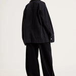 A model shown from the back wearing an oversized black denim jacket and matching black wide-leg trousers, photographed against a plain light gray studio background.