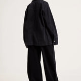 A model shown from the back wearing an oversized black denim jacket and matching black wide-leg trousers, photographed against a plain light gray studio background.
