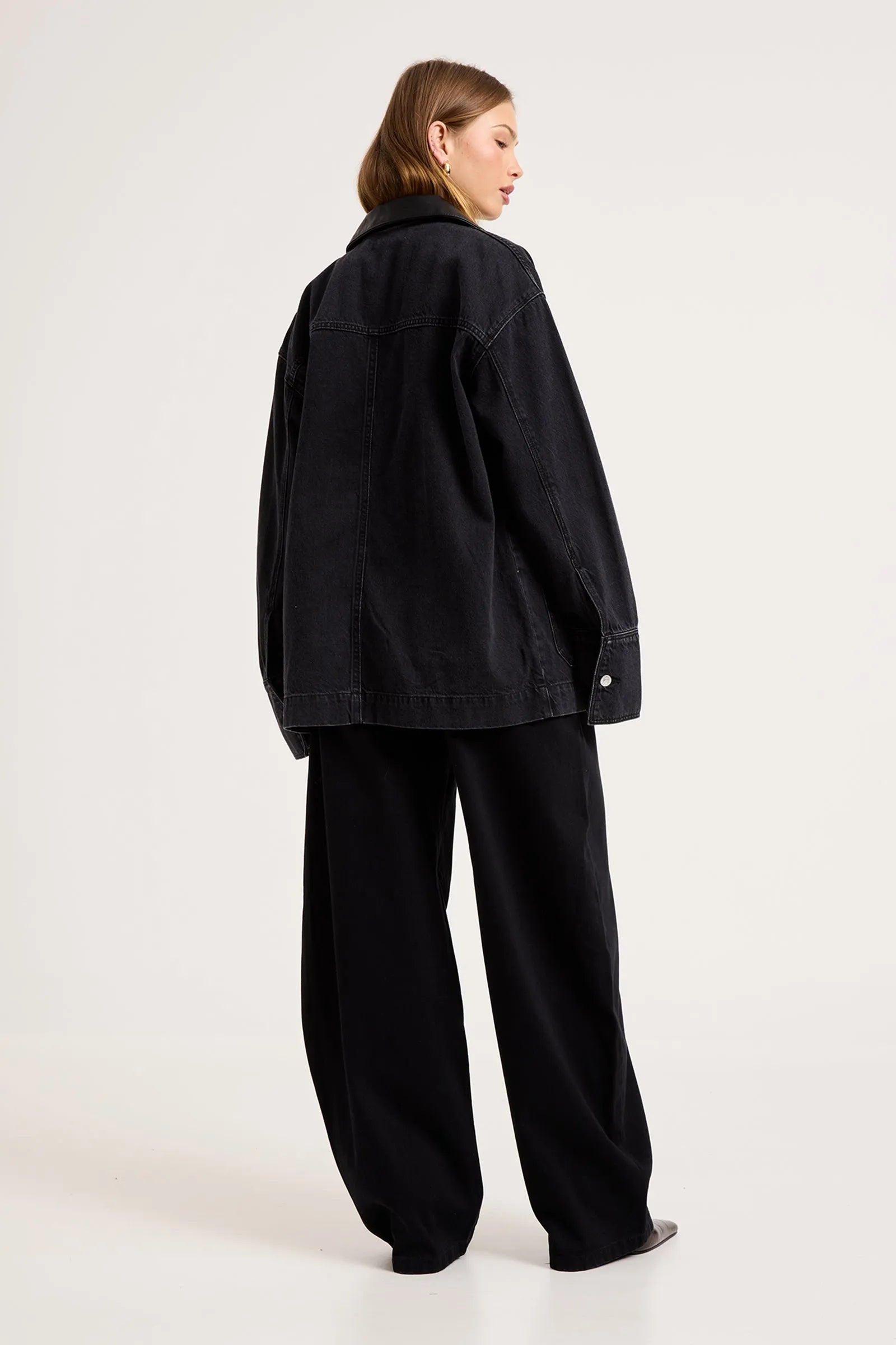 A model shown from the back wearing an oversized black denim jacket and matching black wide-leg trousers, photographed against a plain light gray studio background.