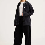 A woman modeling an oversized black denim jacket over a white top and black wide-leg trousers, standing against a plain light gray studio background.