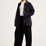 A woman modeling an oversized black denim jacket over a white top and black wide-leg trousers, standing against a plain light gray studio background.