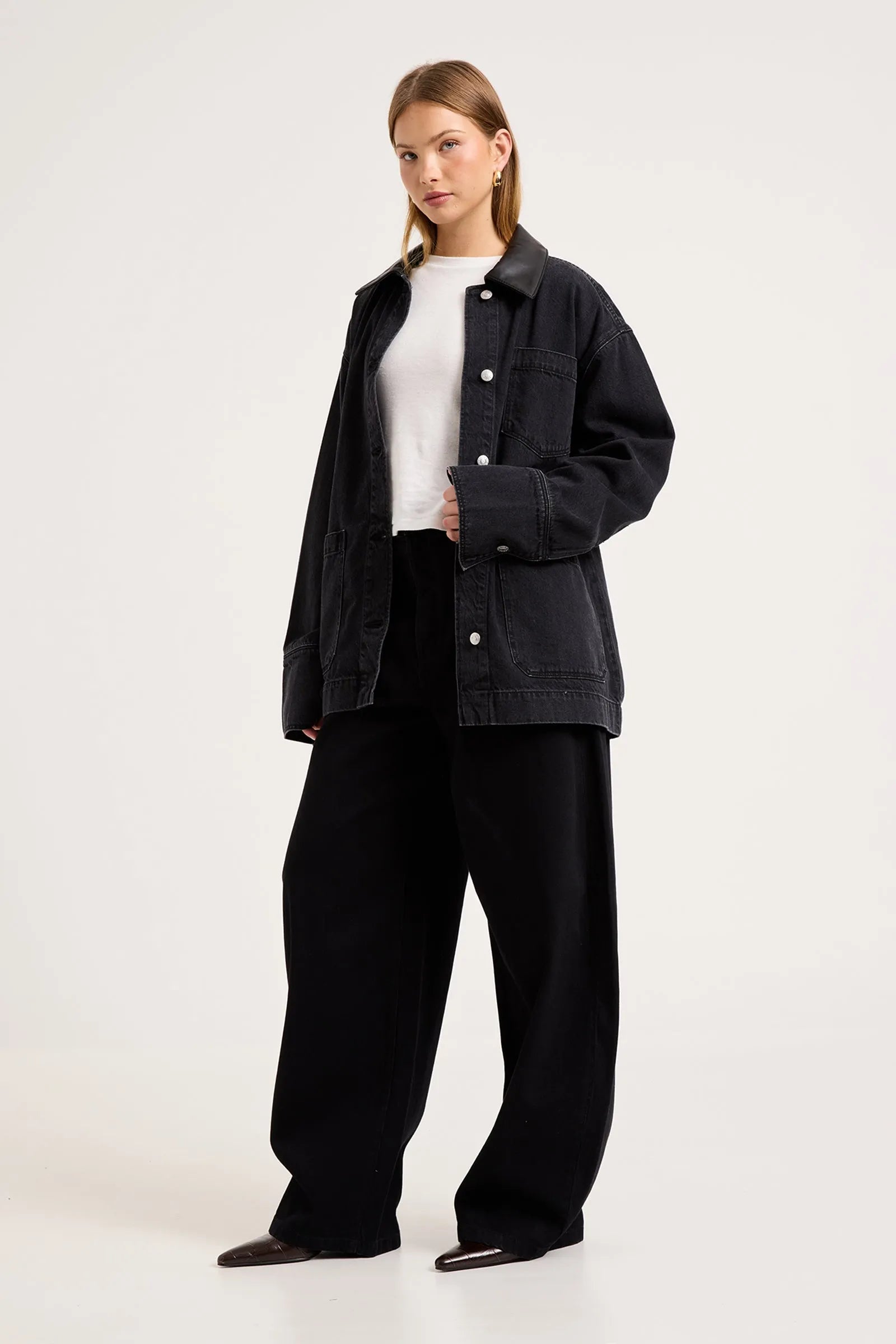 A woman modeling an oversized black denim jacket over a white top and black wide-leg trousers, standing against a plain light gray studio background.