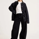A model wearing a black denim jacket over a white t-shirt and black wide-leg trousers, standing against a plain light gray studio background.