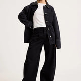 A model wearing a black denim jacket over a white t-shirt and black wide-leg trousers, standing against a plain light gray studio background.