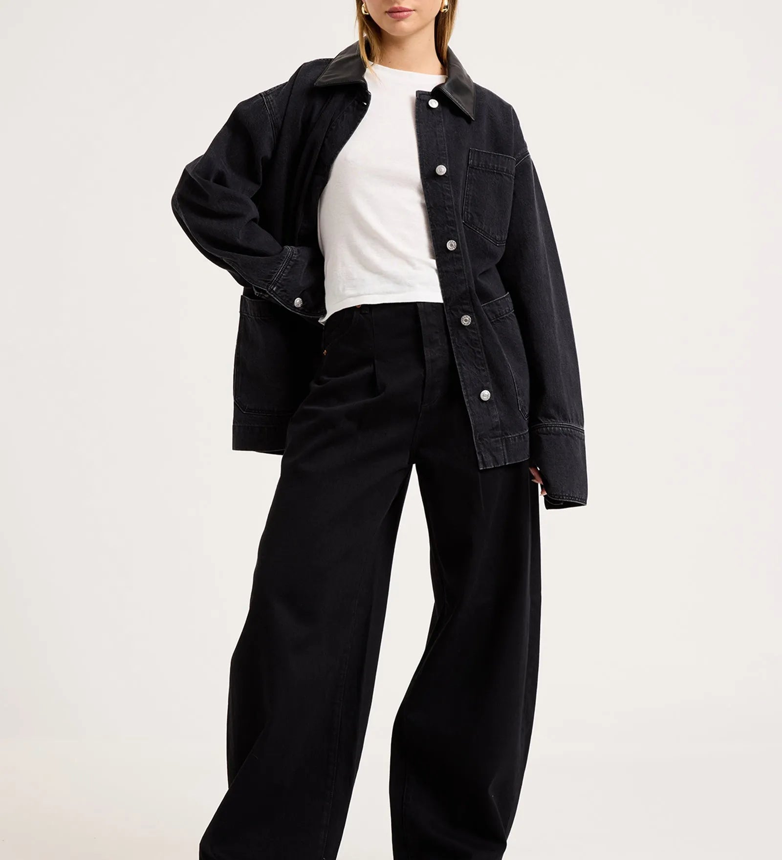 A model wearing a black denim jacket over a white t-shirt and black wide-leg trousers, standing against a plain light gray studio background.