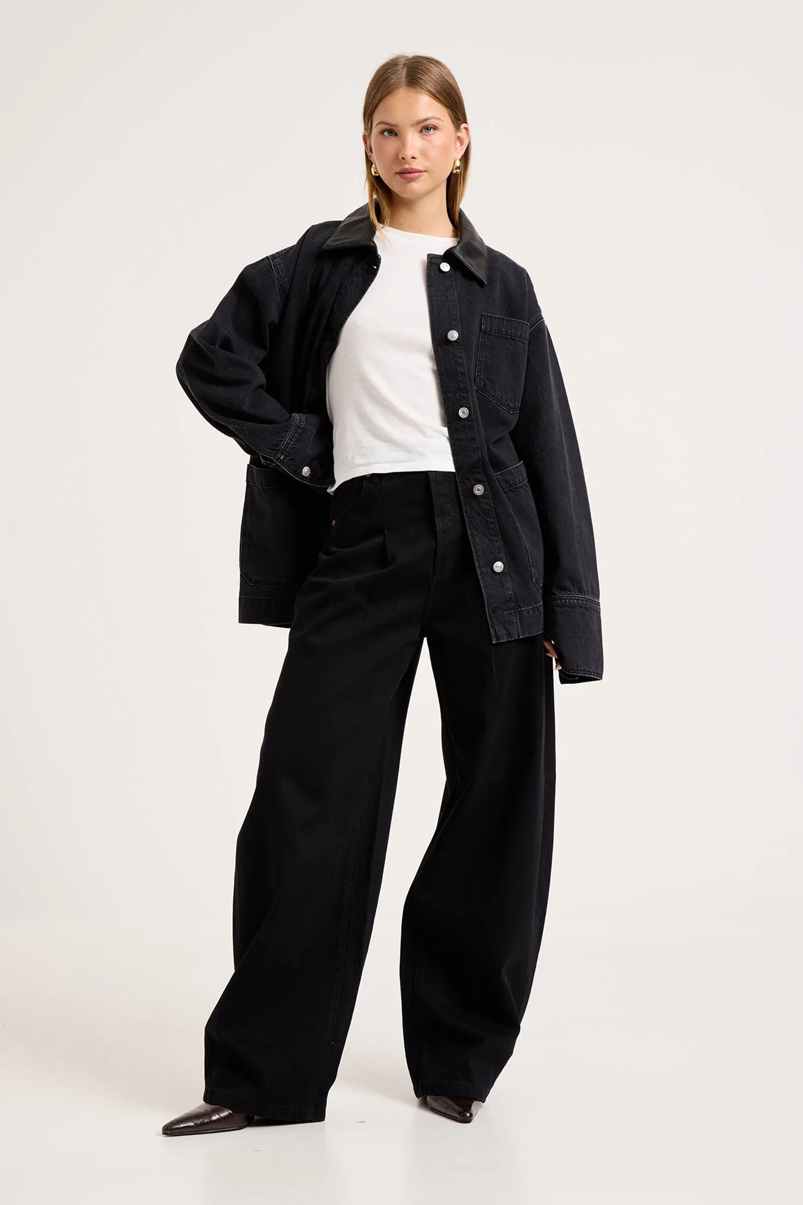 A model wearing a black denim jacket over a white t-shirt and black wide-leg trousers, standing against a plain light gray studio background.