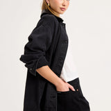 A woman wearing a black denim button-up shirt jacket over a white t-shirt and black trousers, photographed from the side against a light gray studio background.