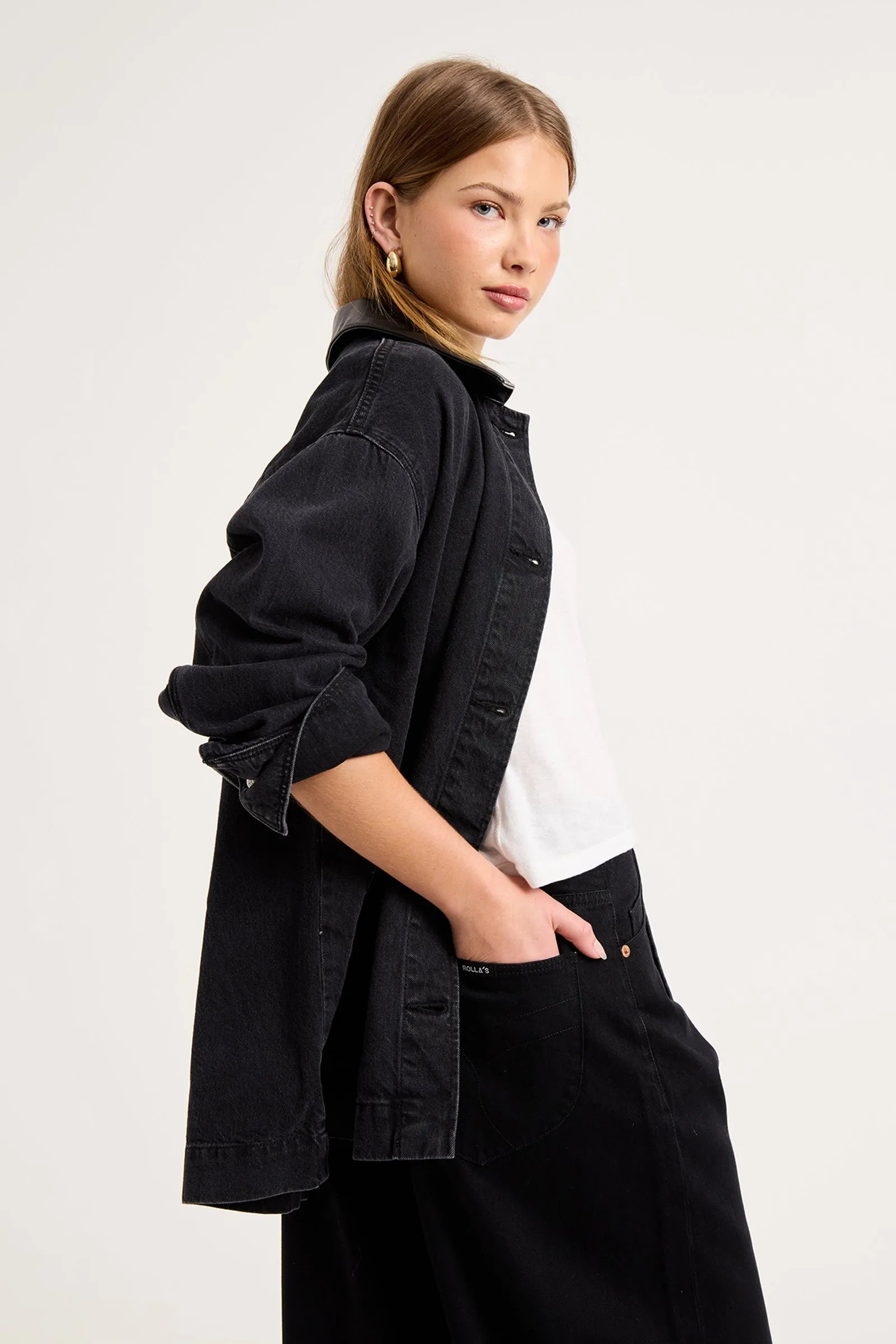 A woman wearing a black denim button-up shirt jacket over a white t-shirt and black trousers, photographed from the side against a light gray studio background.