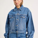 A woman modeling a cropped blue denim jacket with front flap pockets and button closure, paired with matching blue jeans, against a plain light gray studio background.