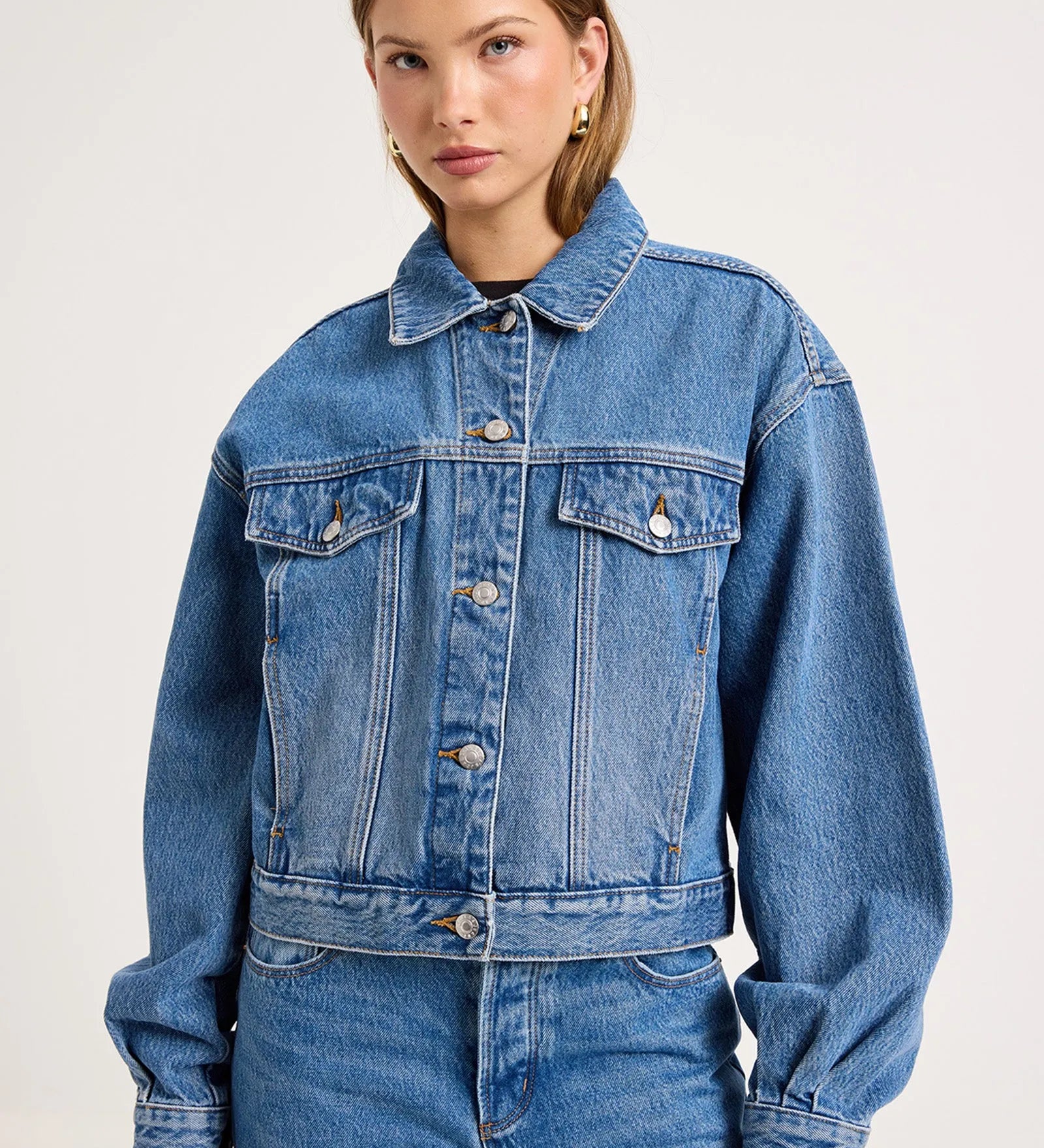 A woman modeling a cropped blue denim jacket with front flap pockets and button closure, paired with matching blue jeans, against a plain light gray studio background.